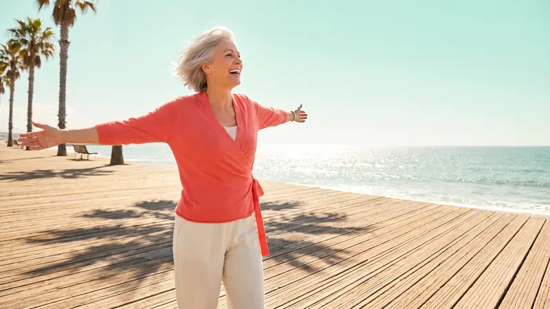 A woman with silver hair on a sunlit boardwalk, arms open, facing the sea.